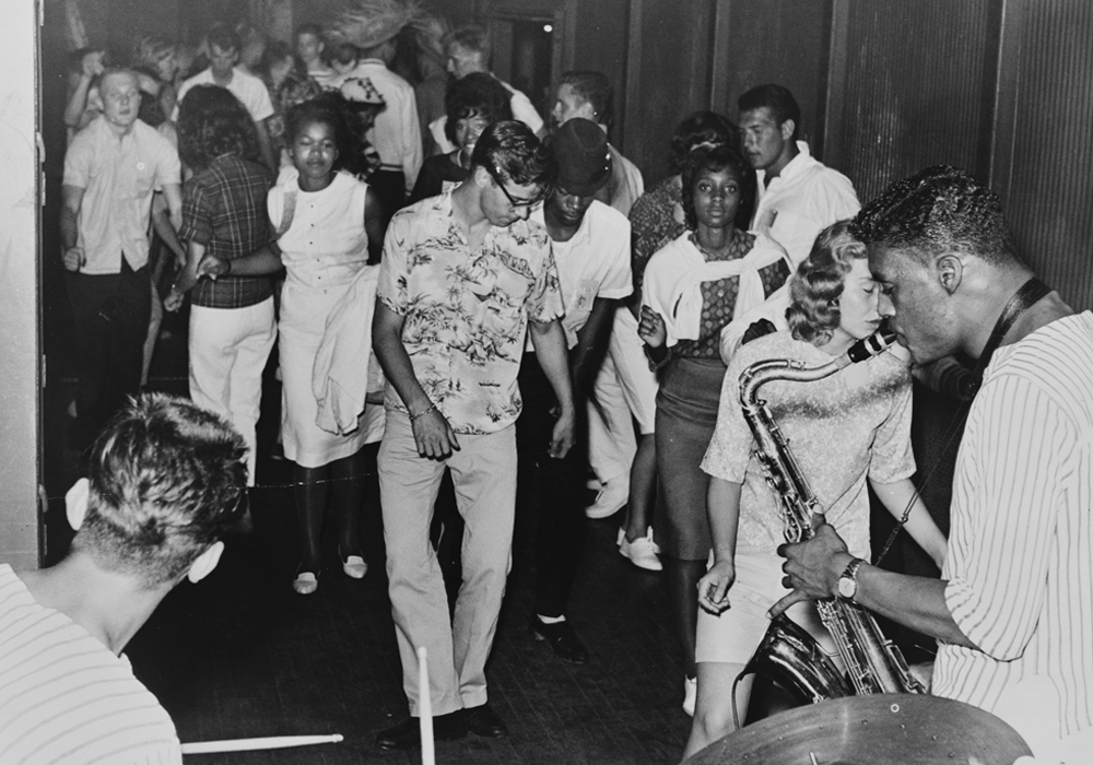 Early 1960s high school students dance as a musician plays a saxophone.