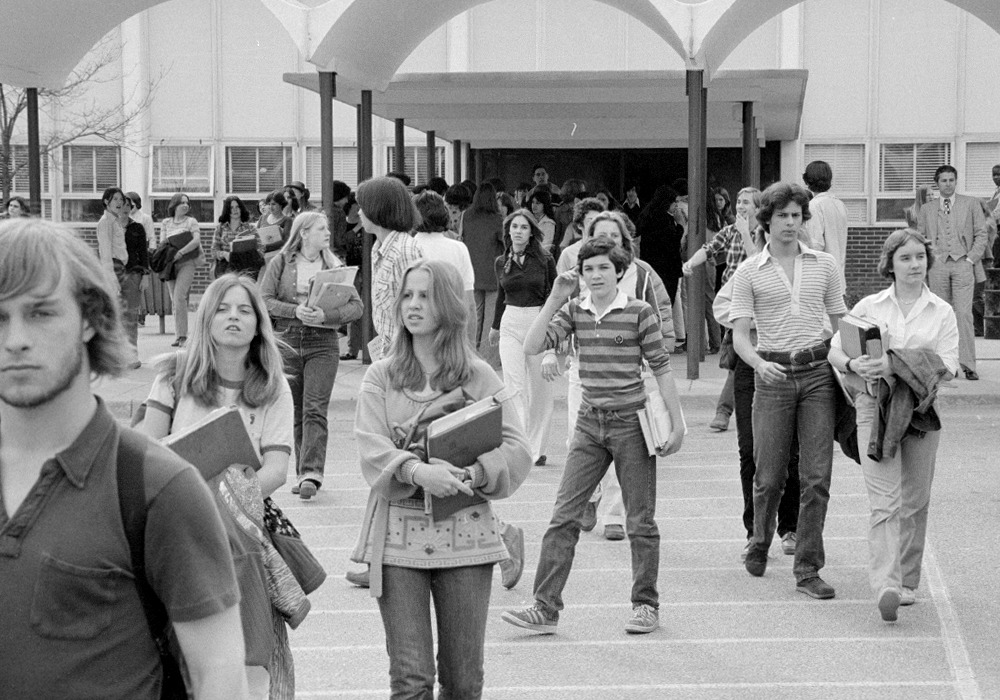 Black and white photo of 1970s students holding books and jackets as they leave a school.