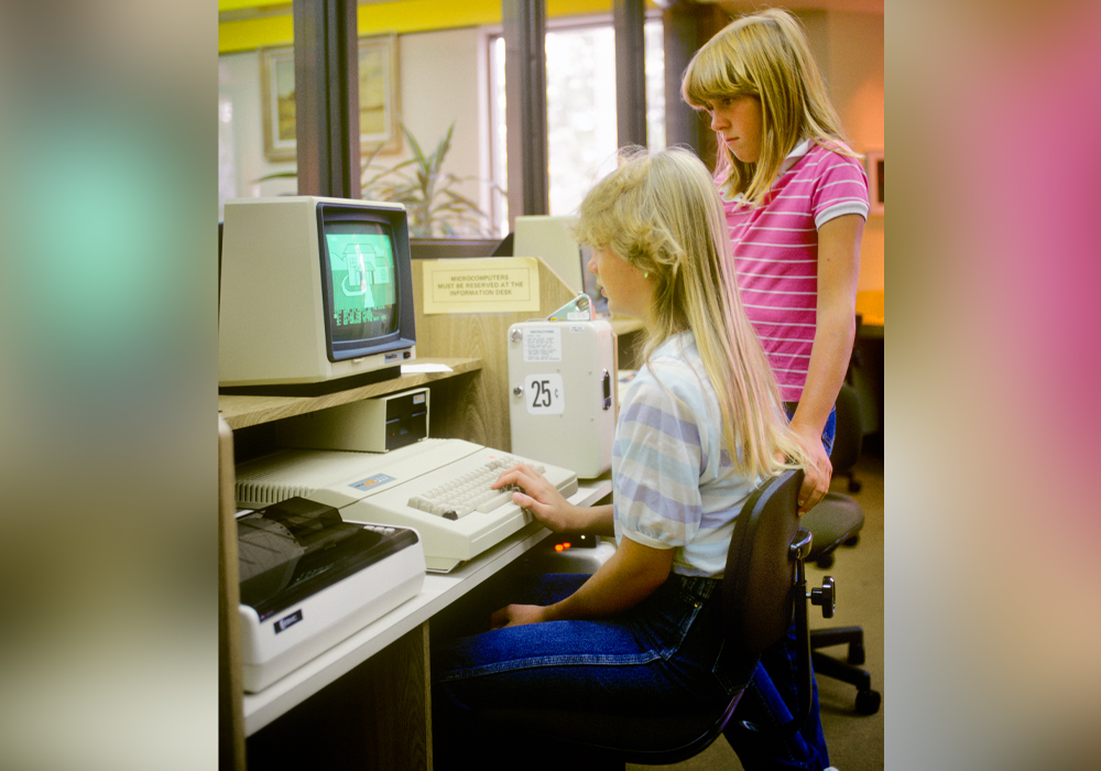Two teen girls work at a 1980s desktop computer.