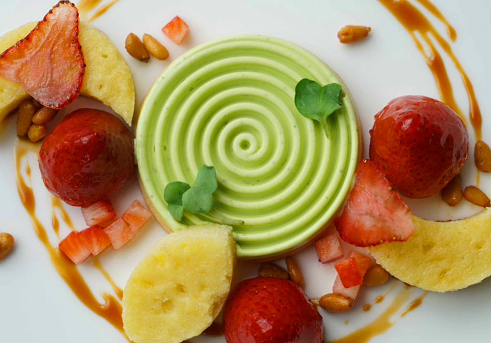 A swirl of green ice cream is on a plate and garnished with basil leaves and some fruit.