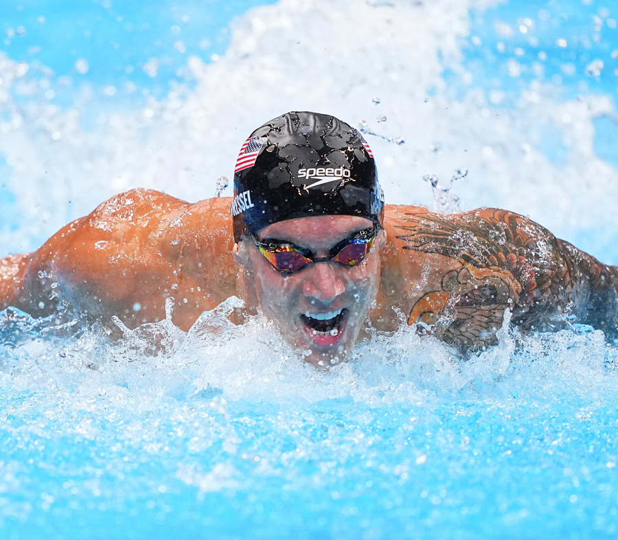 Caeleb Dressel wears a USA swim cap and goggles and does the butterfly stroke in a pool.