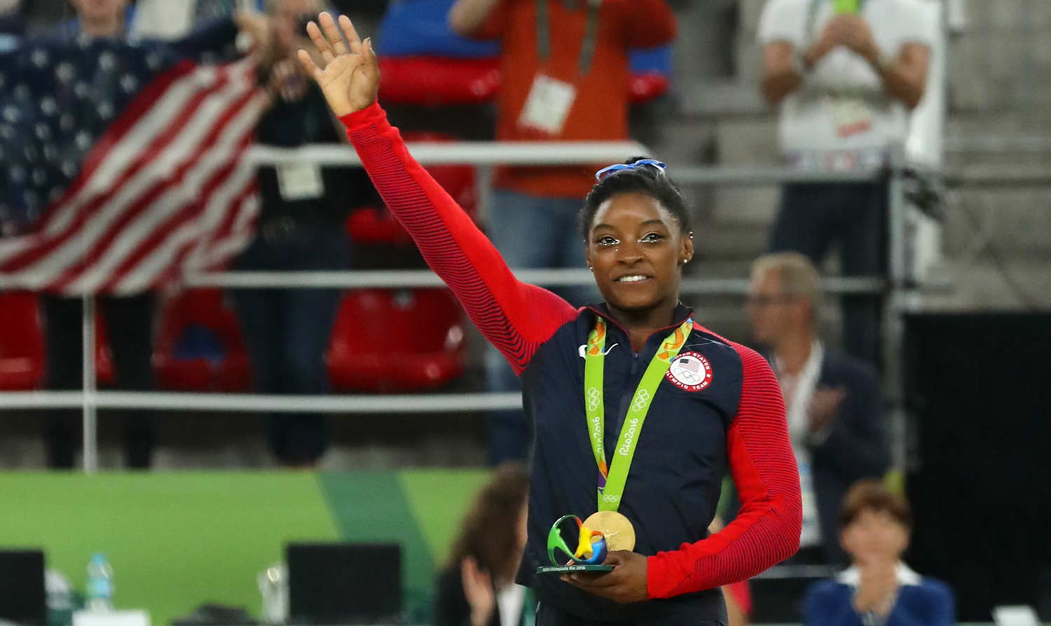 Simone Biles smiles and waves while wearing a USA track jacket with a gold medal around her neck.