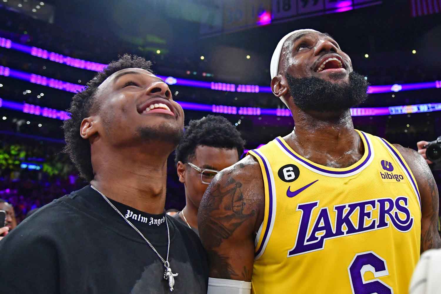 LeBron James in a Lakers uniform and Bronny James stand together in an arena, smiling and looking up.