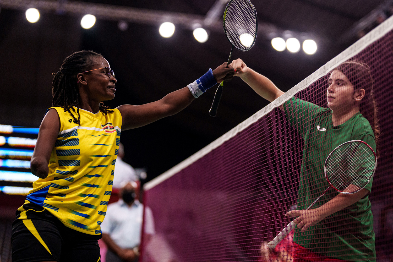 Two athletes on opposite sides of the badminton net bump fists as they hold rackets.