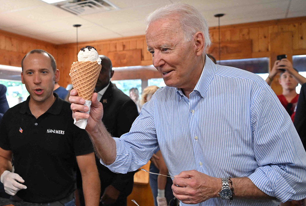 Joe Biden stands in an ice cream store with a few other people holding a waffle cone with vanilla ice cream topped with chocolate chips.