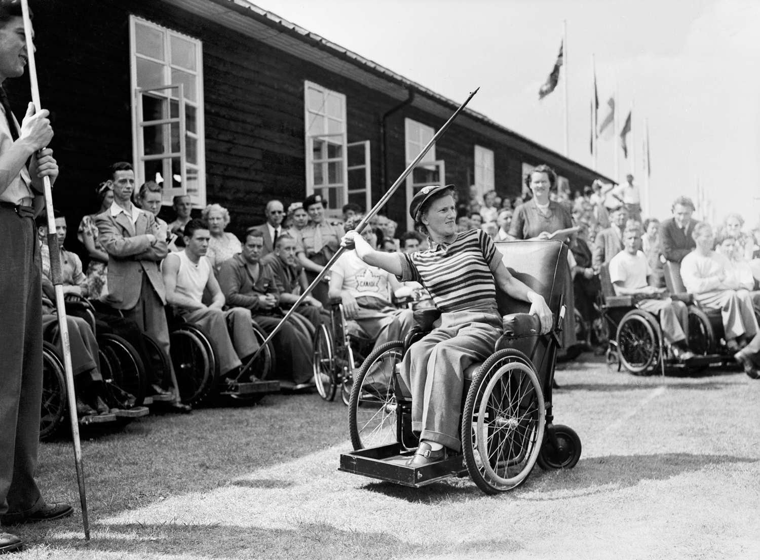 Margaret Webb throws a javelin from her wheelchair as a crowd of people, many in wheelchairs, watches.