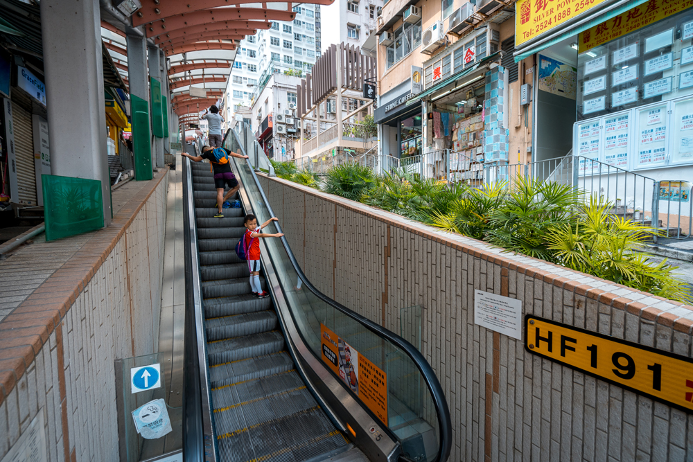 A child and an adult ride an outdoor escalator next to some buildings.