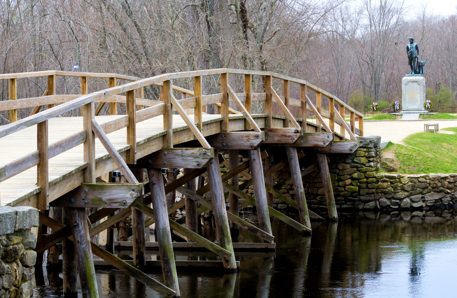 A wooden bridge spans a narrow body of water and a statue of an American Revolution soldier is in the background.