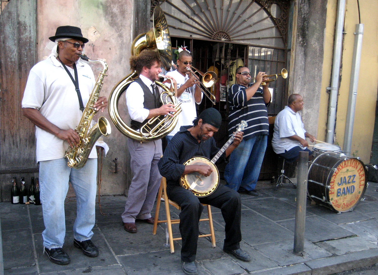 Six musicians who make up the Preservation Hall Jazz band perform outdoors with horns, a drum, and a banjo.