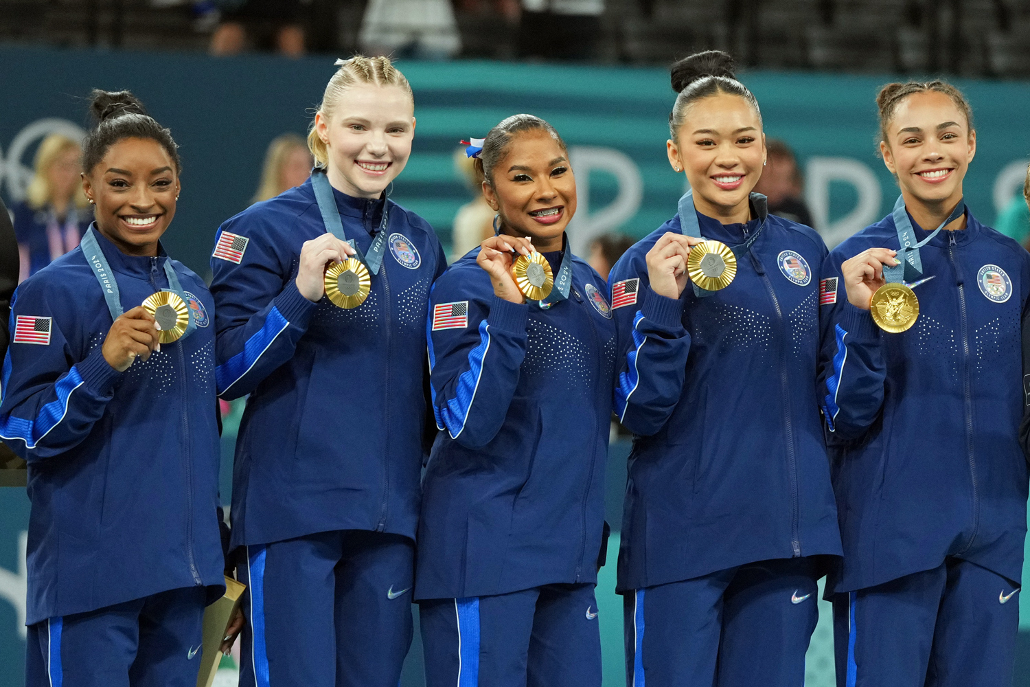 The five members of the US women’s gymnastics team pose wearing warm up suits and gold medals.