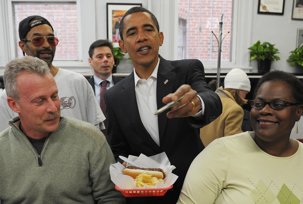 Barack Obama stands in a store with a few other people and gives cash for the chili dog he is holding.