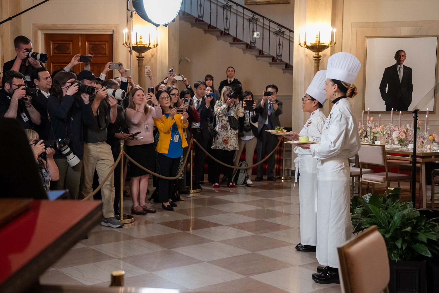 Cristeta Comerford and another chef stand in a White House room holding plates as a group of photographers take pictures.