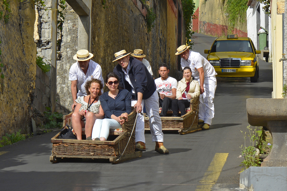 Two wicker toboggans have two passengers each and are each being pushed by two men.