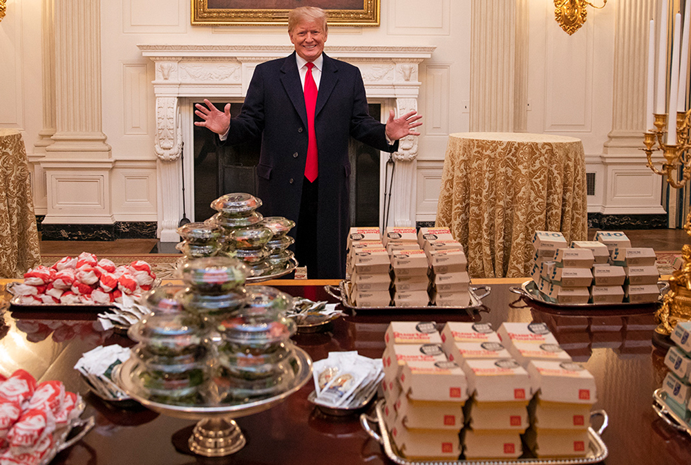 Donald Trump stands behind a table filled with burgers and other fast food items in their packaging.