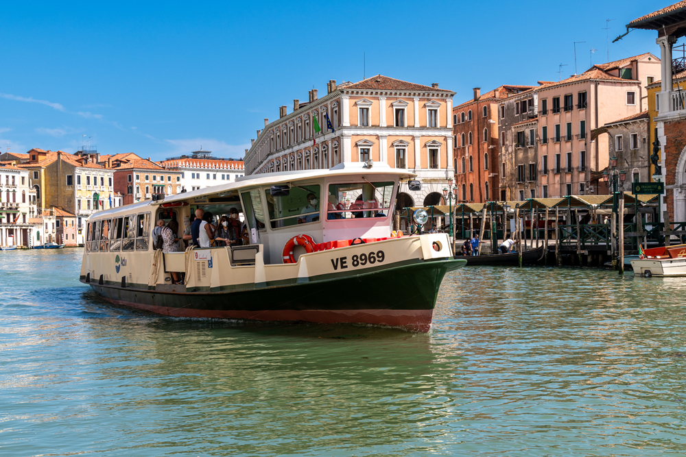 Several passengers are onboard a vaporetto on a canal that is lined with old buildings.