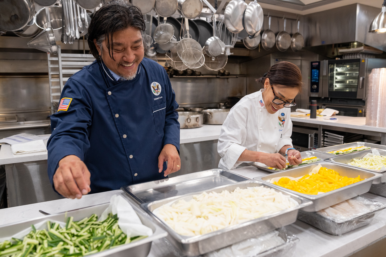 Edward Lee wears a blue chef’s coat and Cristeta Comerford wears a white chef’s coat as they stand at a counter placing food on trays.