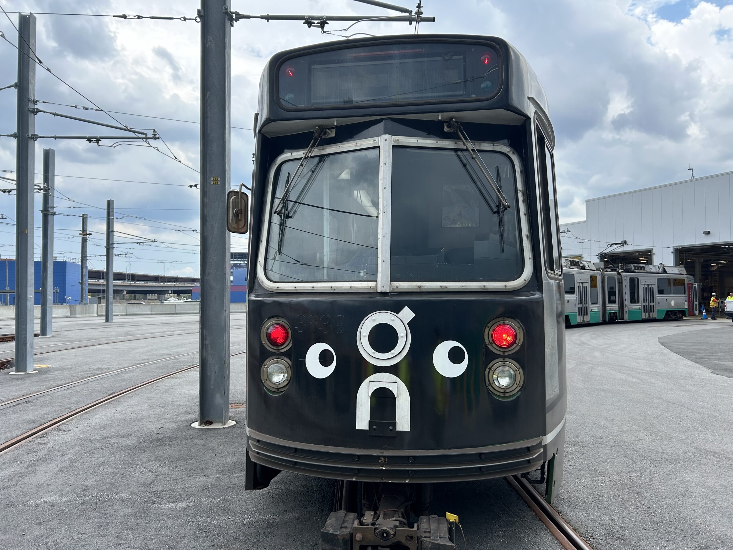 A black subway car with decal eyes on the front leads a number of other cars on an outdoor track.