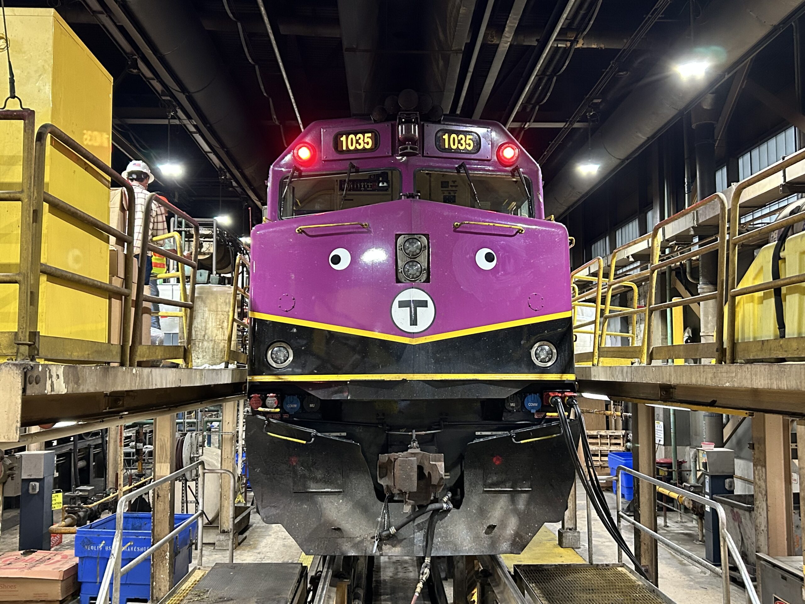 The front of a purple subway car with decal eyes that is sitting on an indoor track.