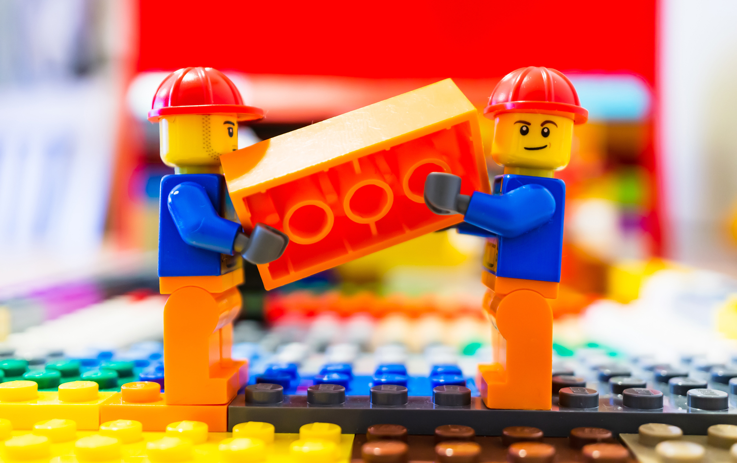 Either end of a Lego brick is held by two Lego people wearing hard hats.