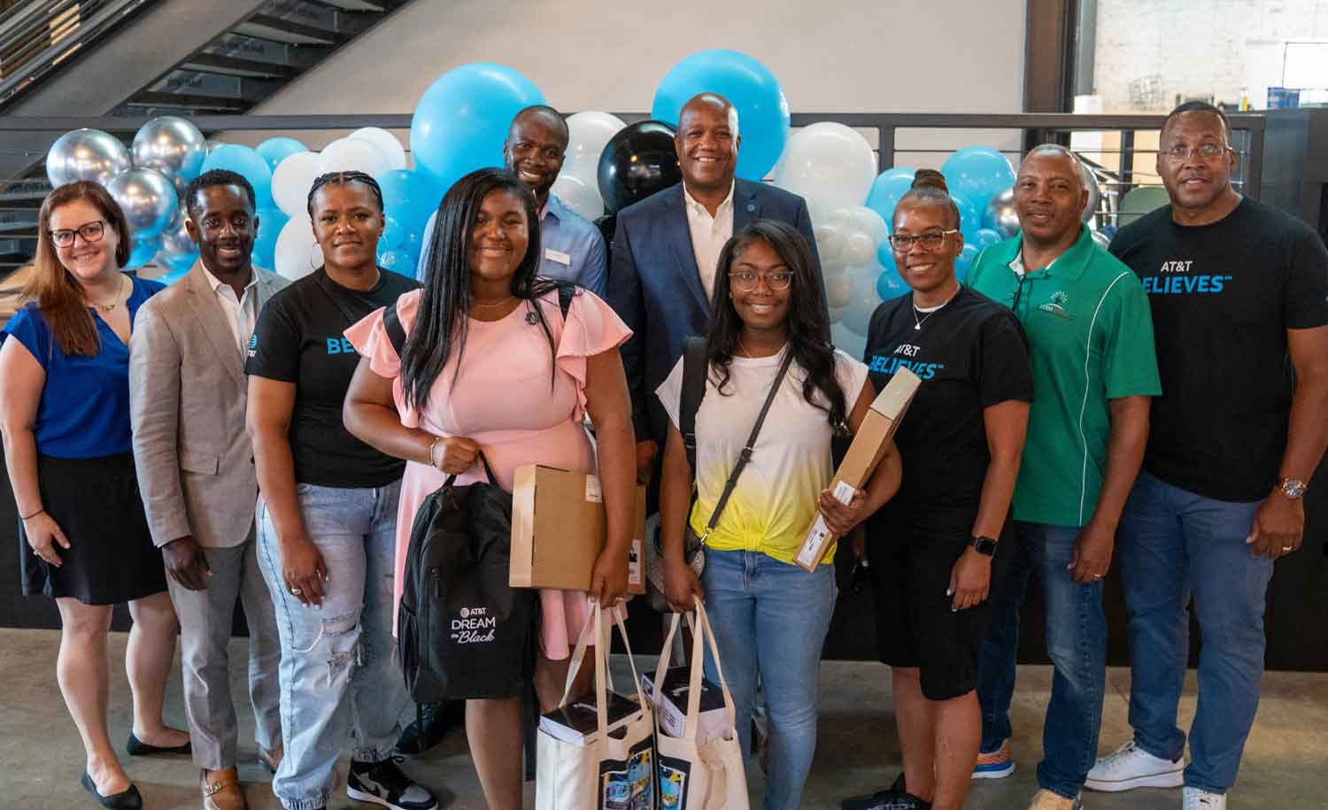 Two teen girls pose with several men and women in front of balloons while holding gift bags.