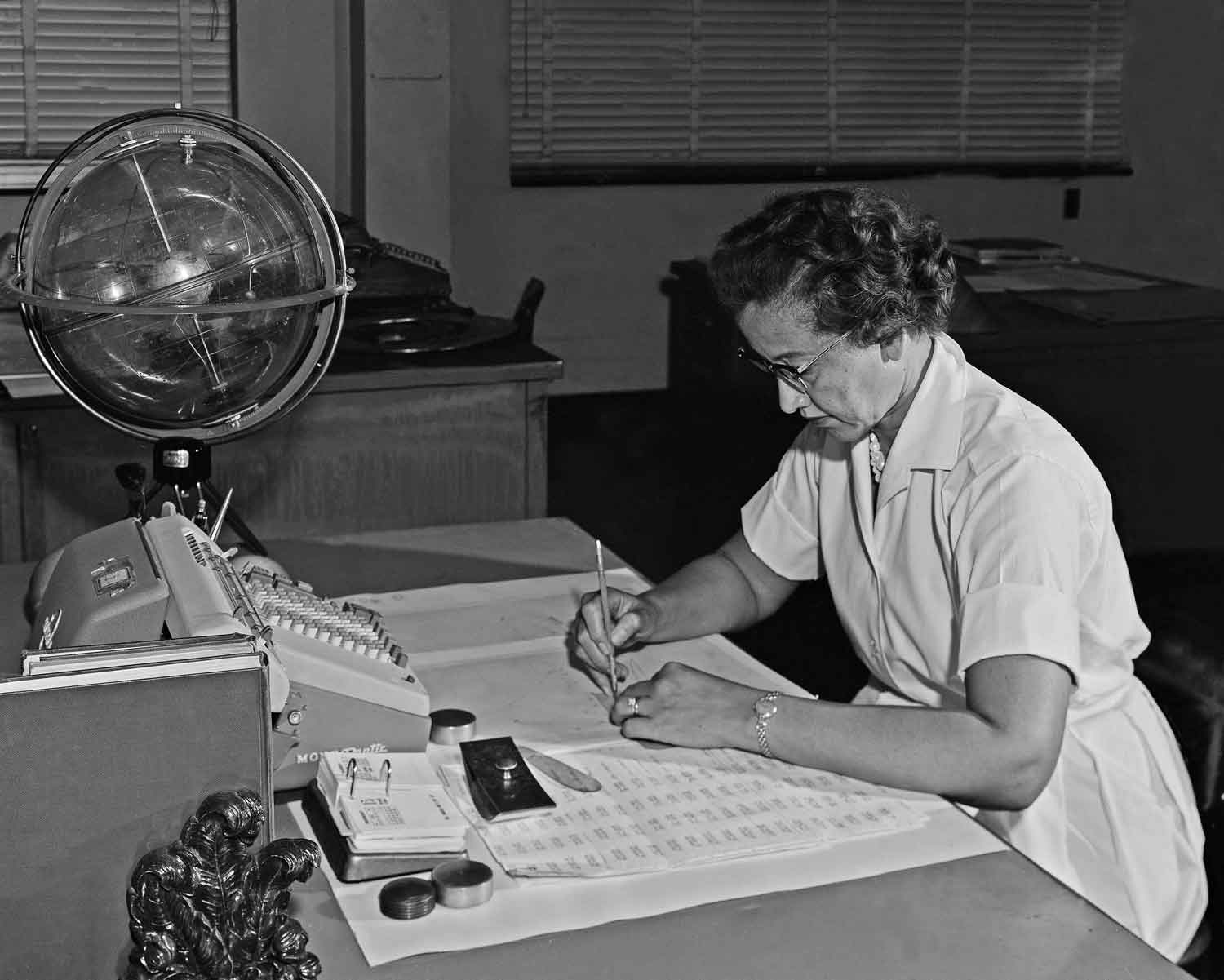 Katherine Johnson writes at a desk that holds an adding machine and several papers.