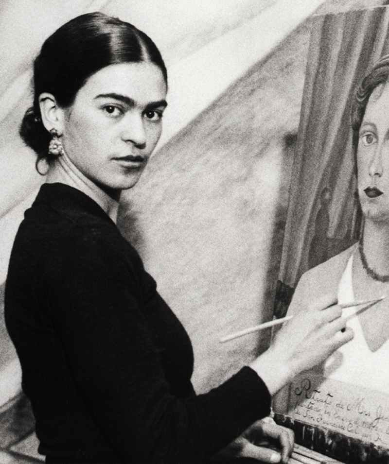 Frida Kahlo looks at the camera as she paints a portrait of a woman.