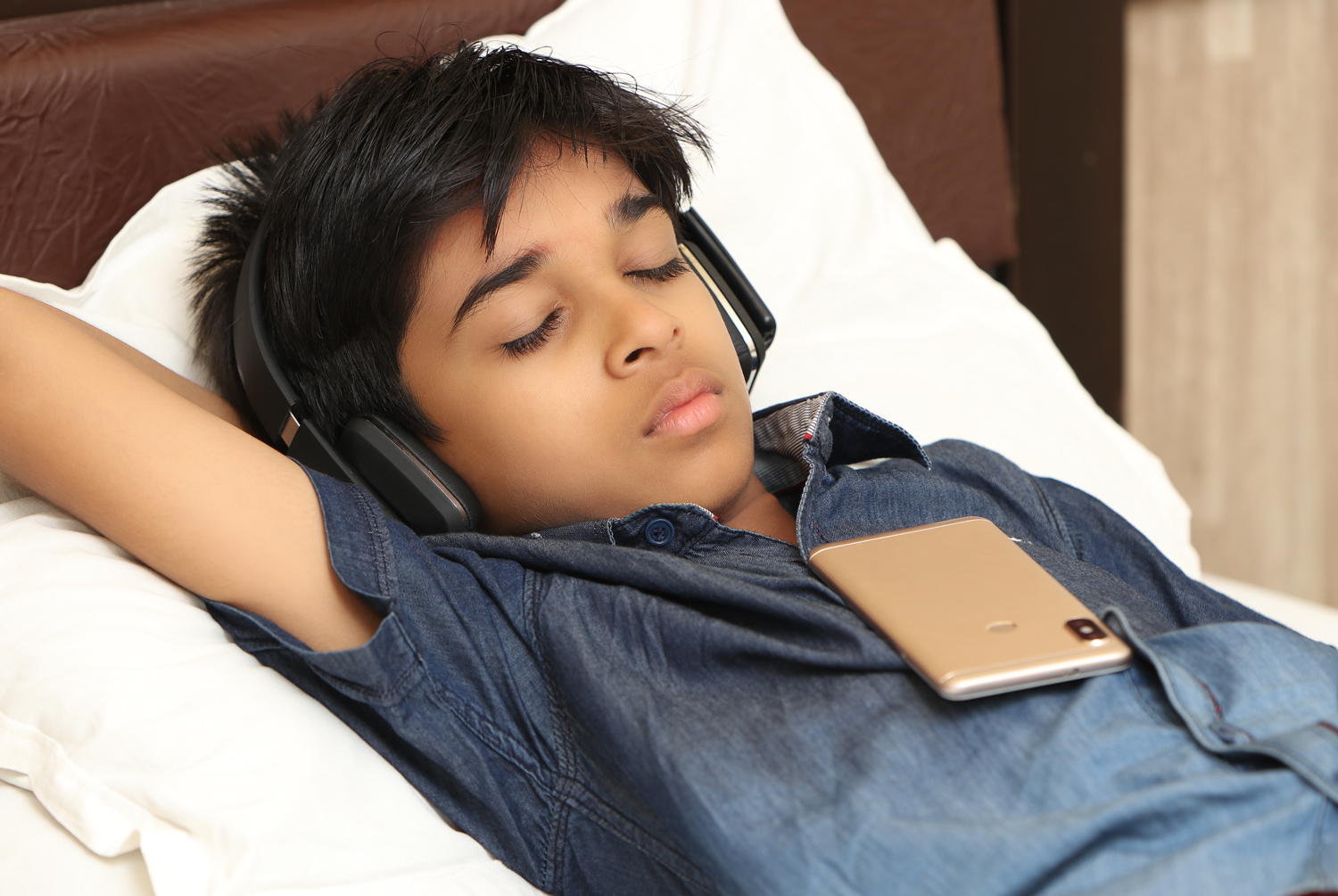 A teenager sleeps on a bed with a phone on his chest and while wearing headphones.