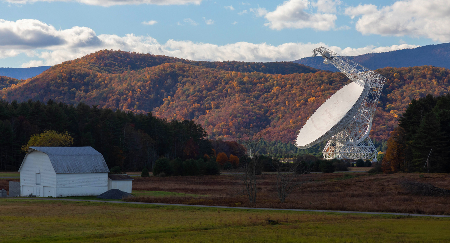 A barn is dwarfed by the large white telescope that sits near it.