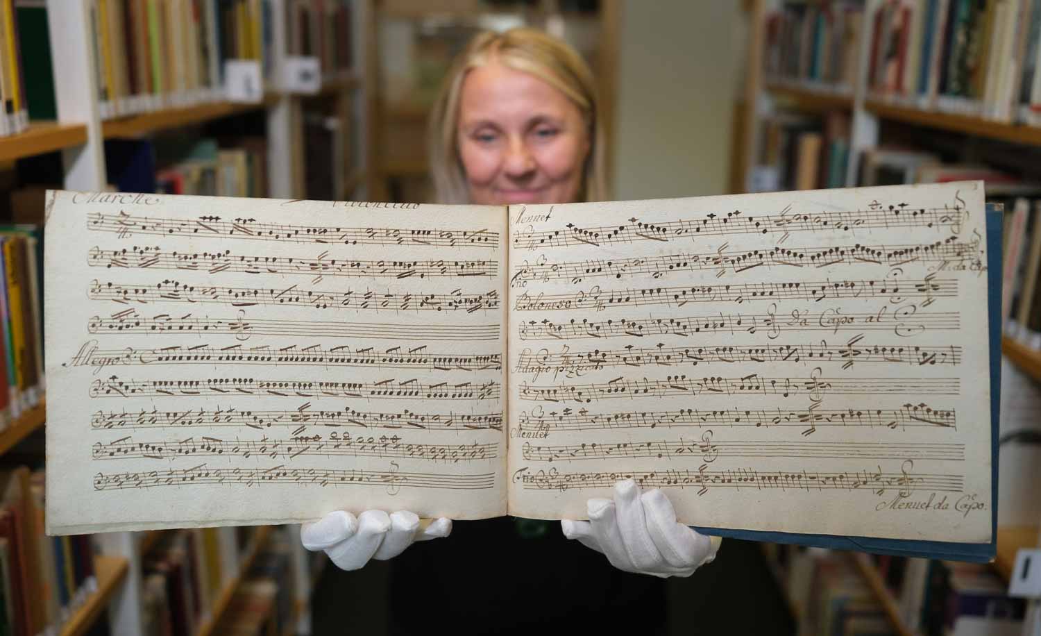 A woman wearing protective gloves holds up two sheets of handwritten music.