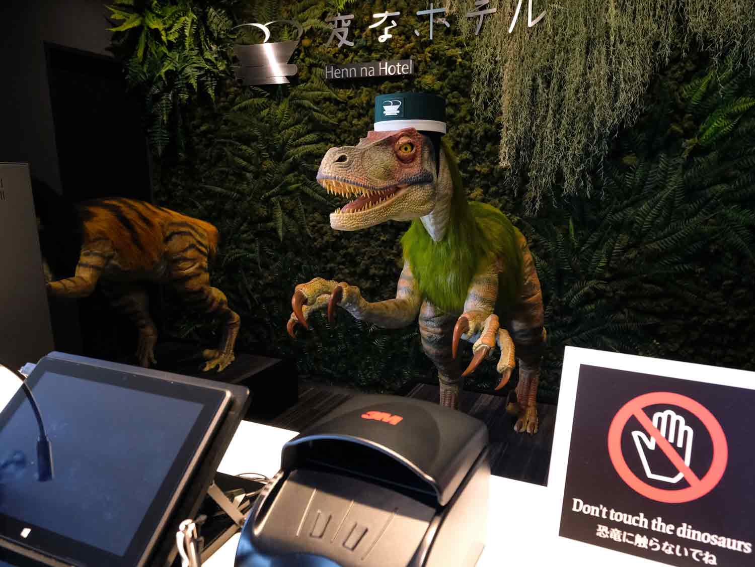 A dinosaur robot wears a uniform and stands behind a hotel reception desk.