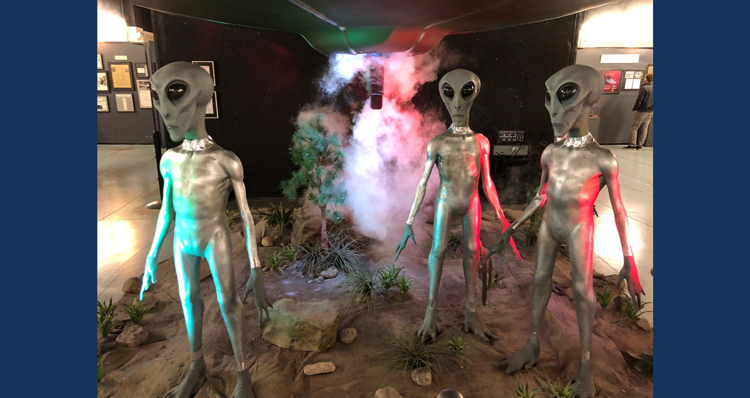 An artist’s conception of three aliens standing on Earth below a lit-up UFO.