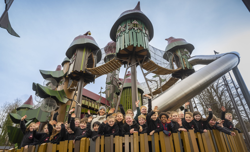 A group of schoolchildren and an adult wave in front of a large play structure with many turrets, bridges, and slides.