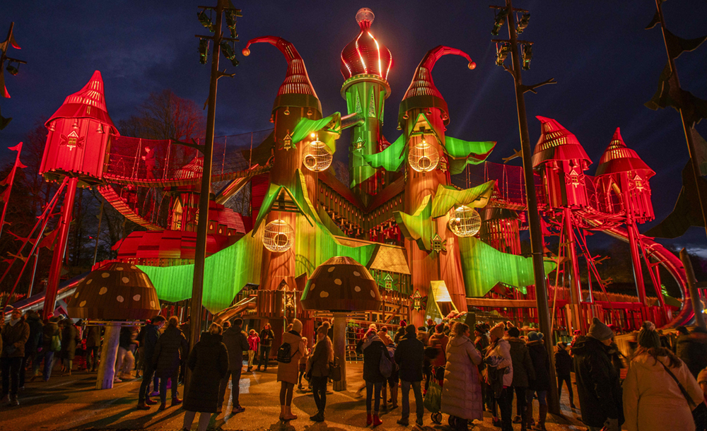 A large play structure with many turrets, bridges, and slides, lit up at night with a crowd of people around it.