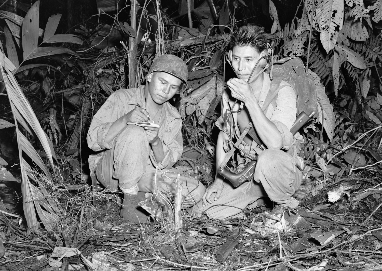 Two soldiers in uniform sit among vegetation as one writes in a notebook and the other holds equipment.