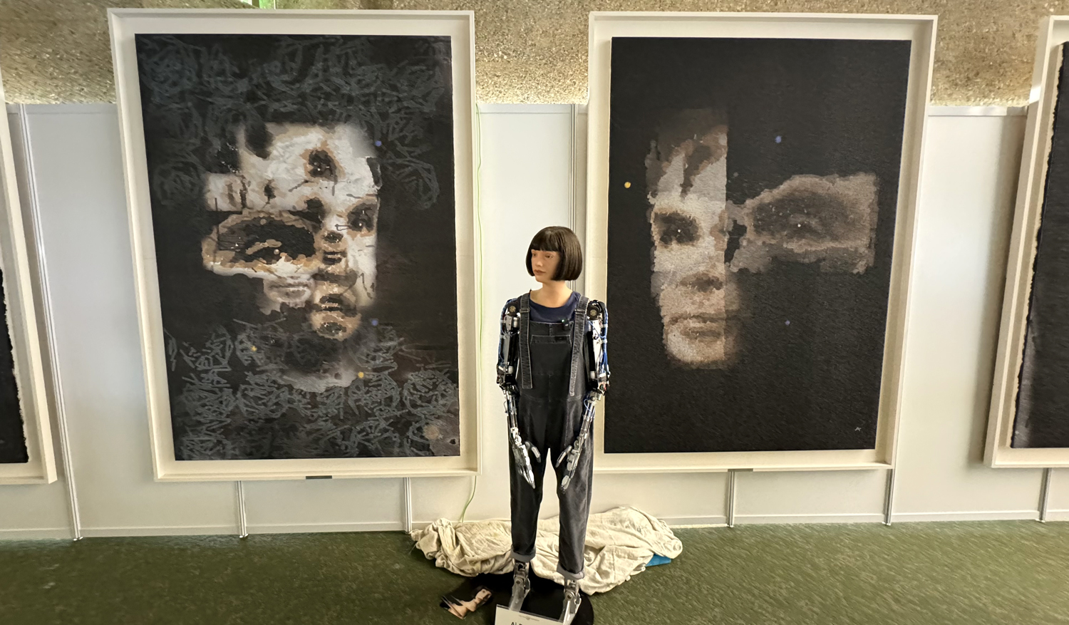 A robot that looks like a woman stands in front of two paintings of Alan Turing.