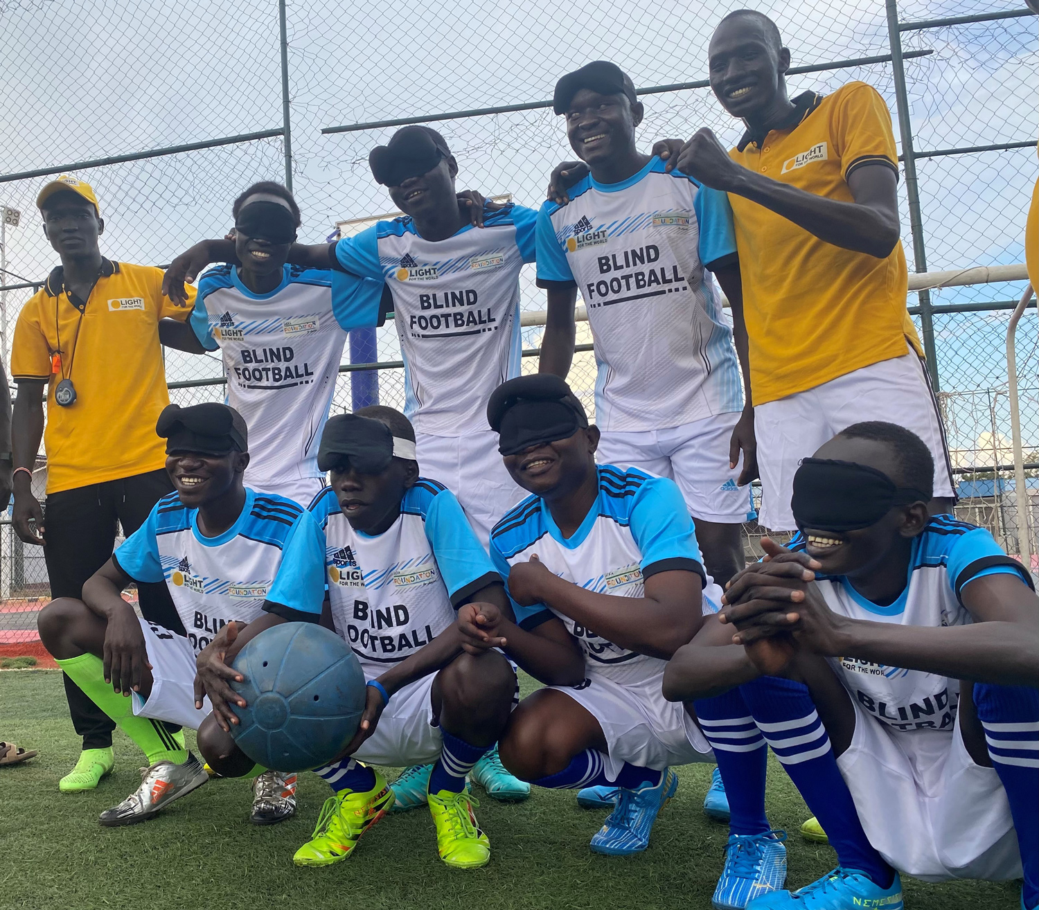 Seven men wearing uniforms reading blind football pose with two coaches in yellow polo shirts.