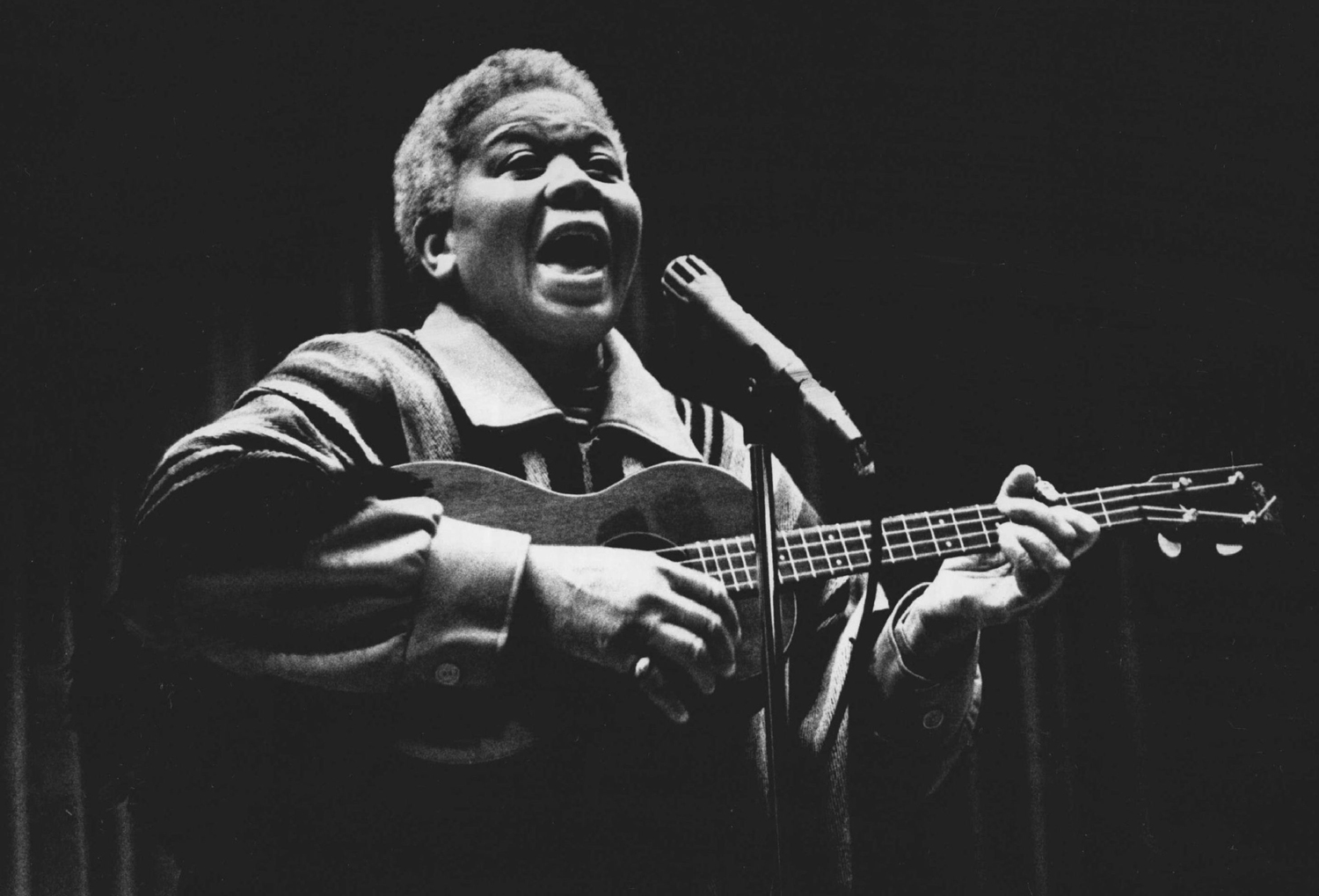 Ella Jenkins plays a ukelele as she sings at a microphone.