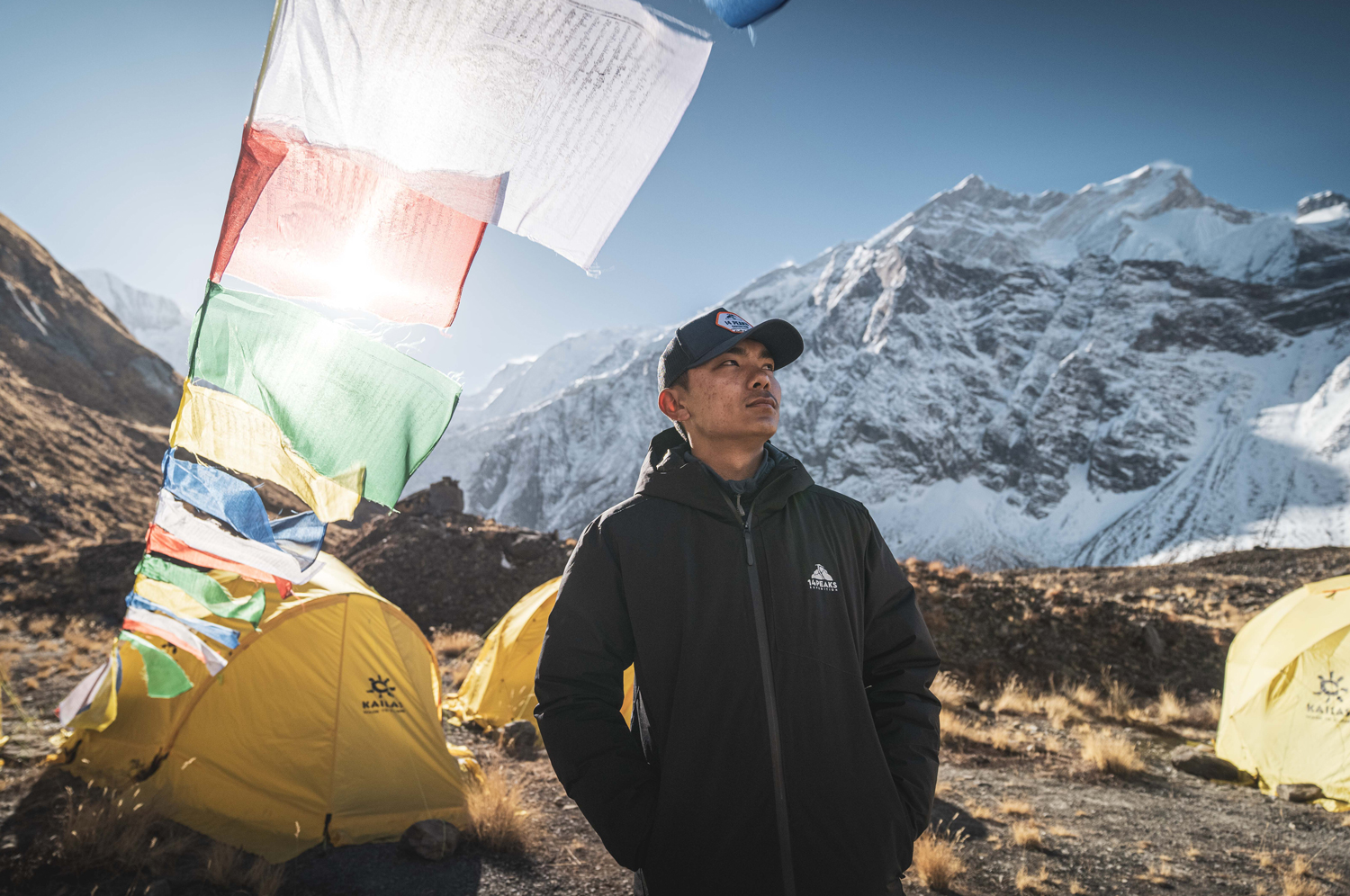 Nima Rinji Sherpa stands in front of a group of yellow tents on a mountainside with a snow-topped mountain in the background.