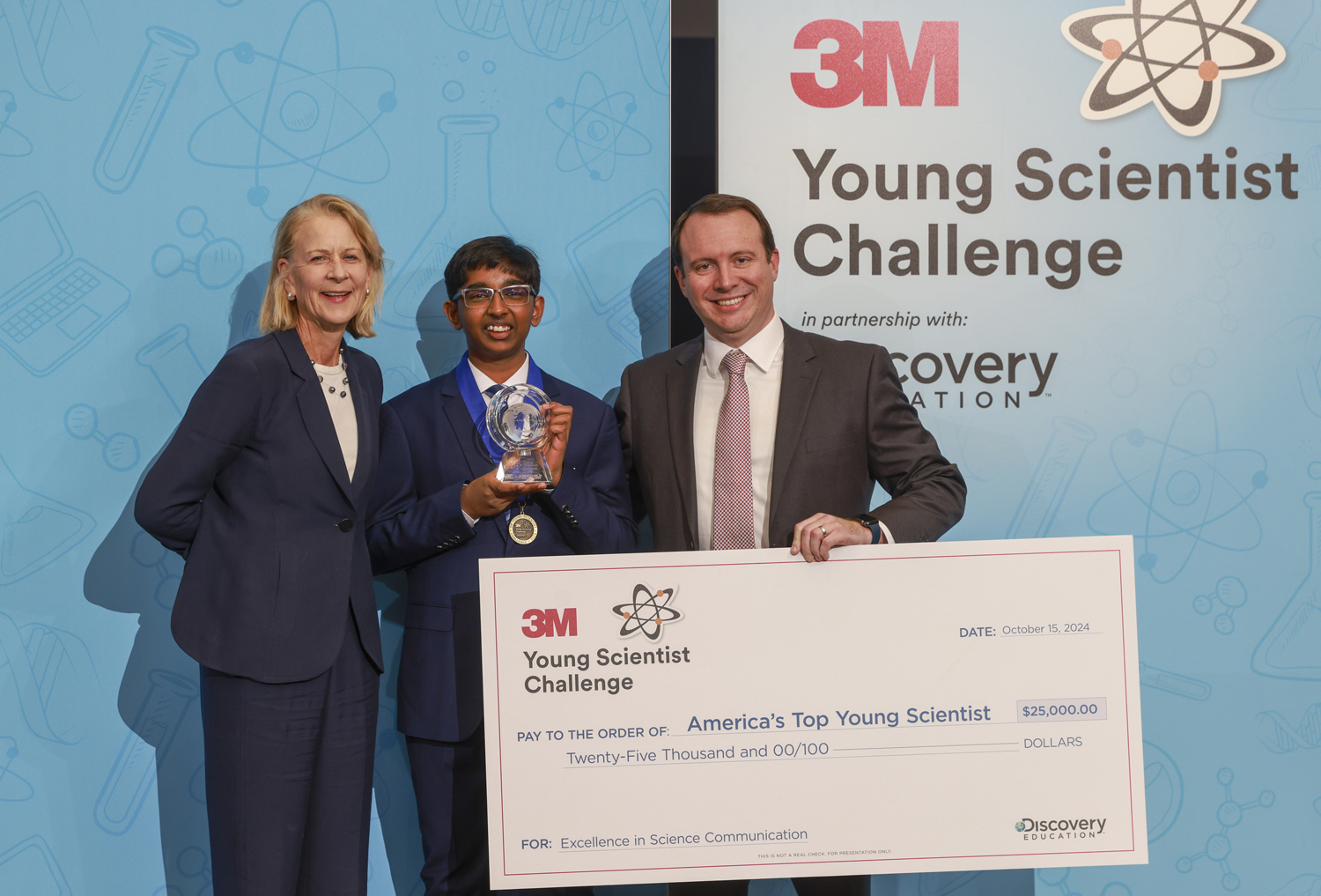 Sirish Subash, a 3M Discovery Education official, and a 3M official pose with a plaque, a medal, and a large check for $25,000.