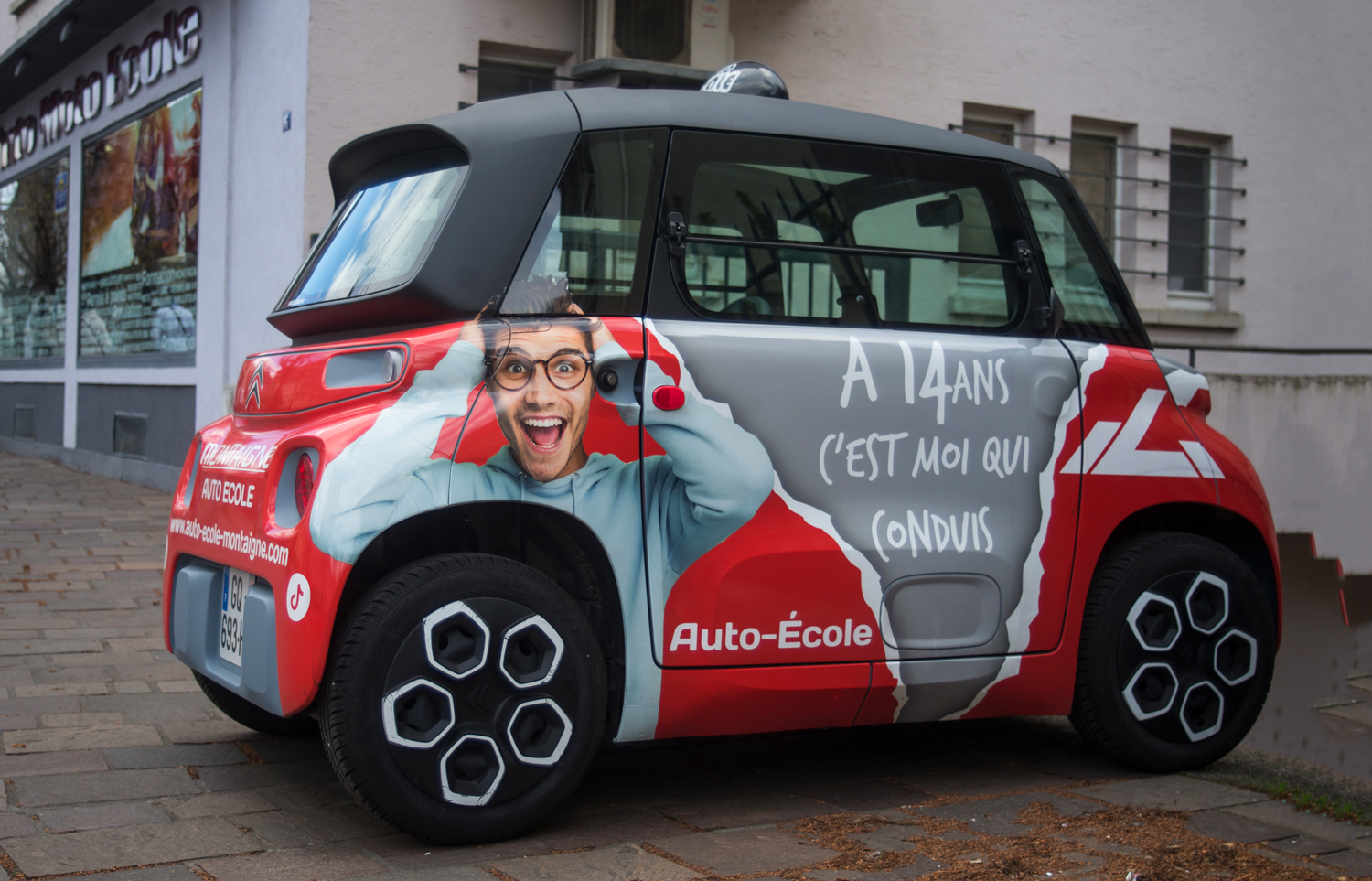 A Citroën Ami car is painted with an image of a teen and the words A 14 ans c’est moi qui conduis.