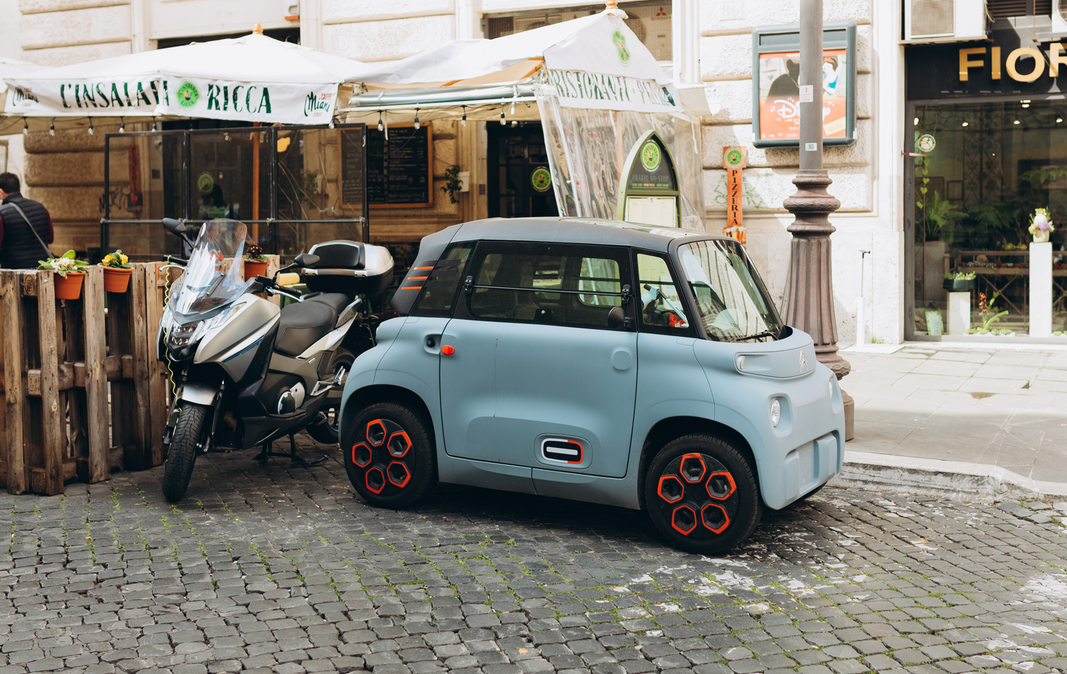 A Citroën Ami is parked next to a motorcycle in front of a restaurant and a storefront.