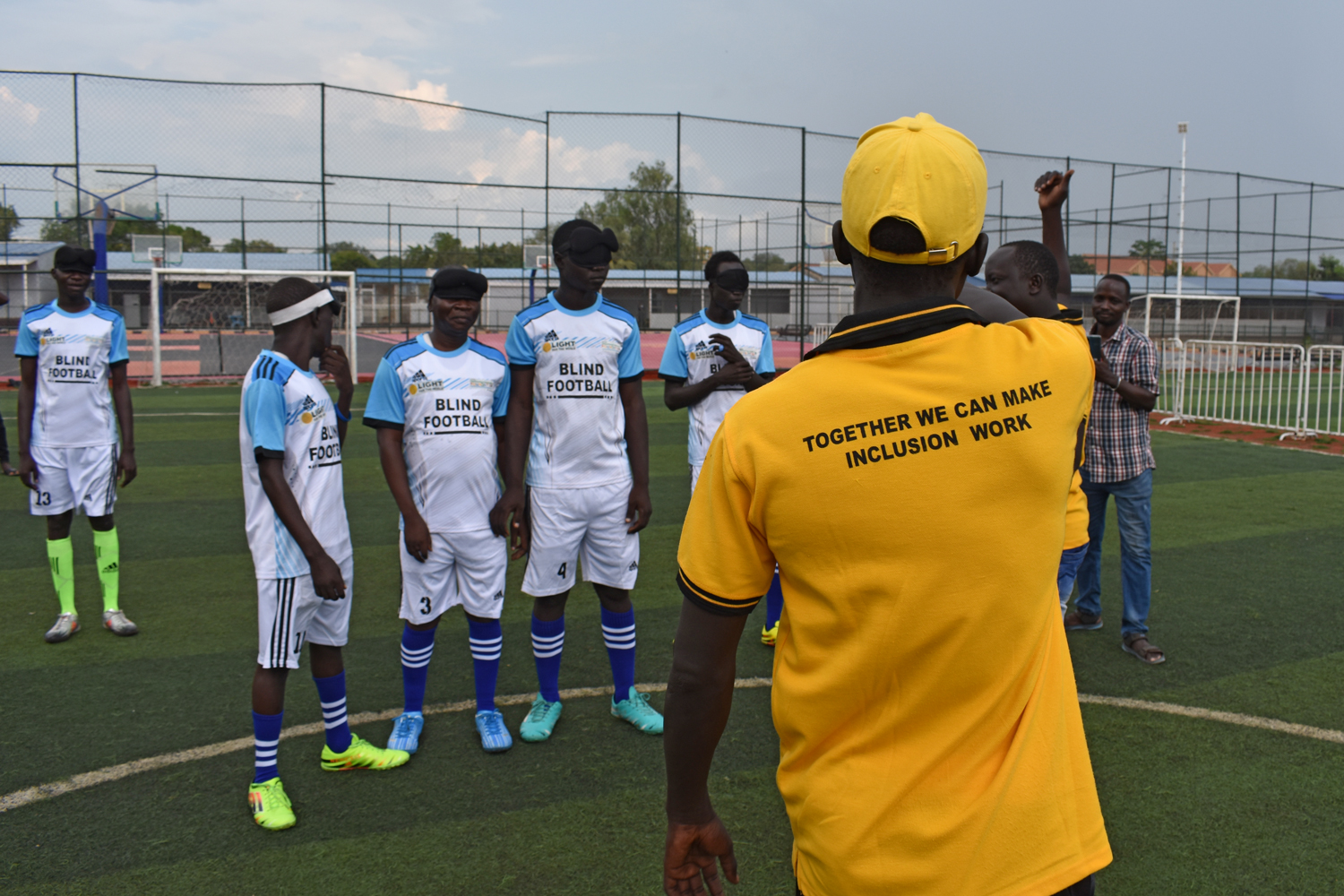 A coach speaks to members of the soccer team while wearing a shirt that reads Together we can make inclusion work.