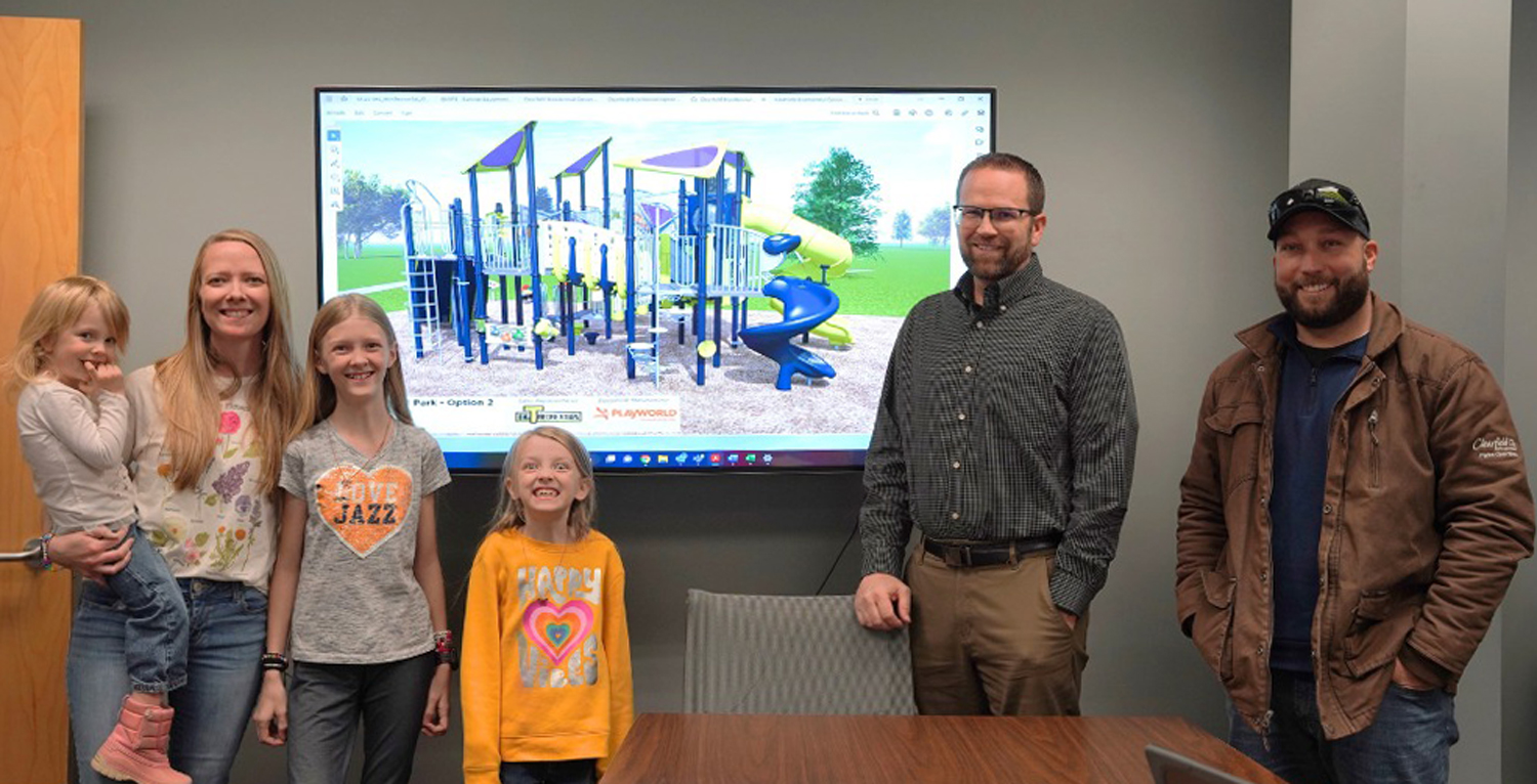 Rosili Olson with her sisters, mother, and Clearfield city officials in a room in front of a rendering of her playground design.