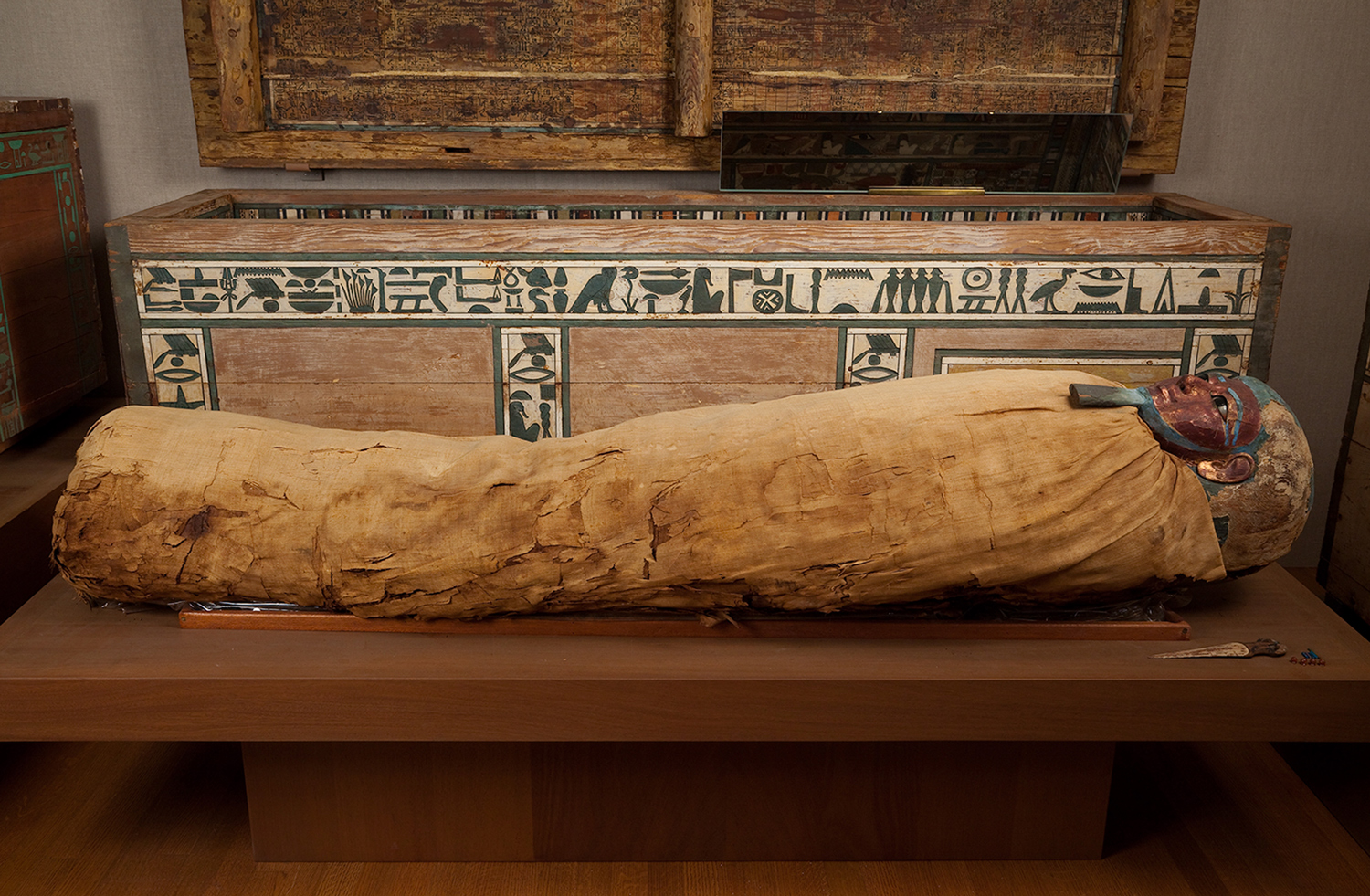 A mummy with a mask on its face lies in front of a decorated sarcophagus.