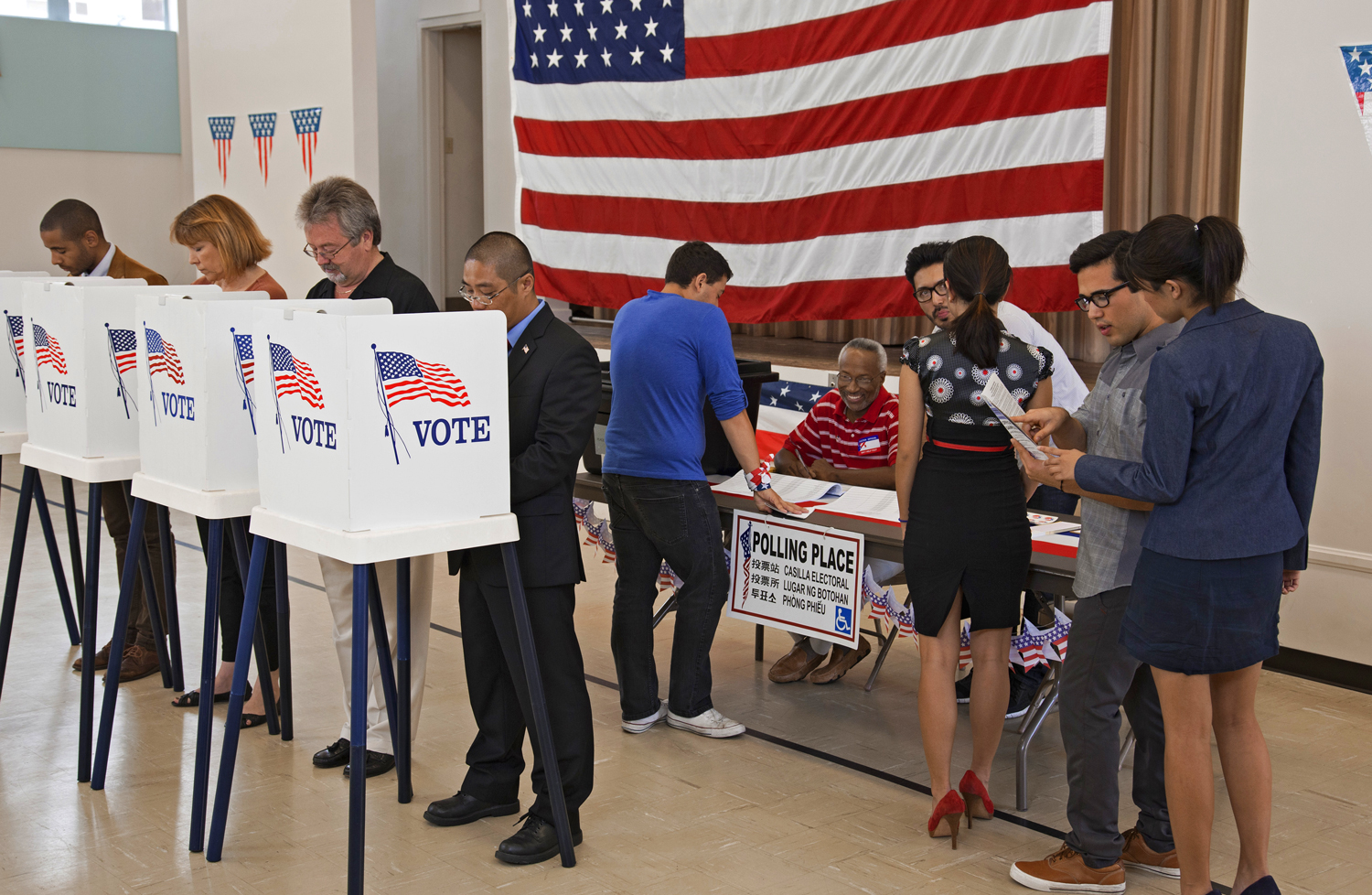 At a polling place, some voters sign in with poll workers while others stand at voting booths.