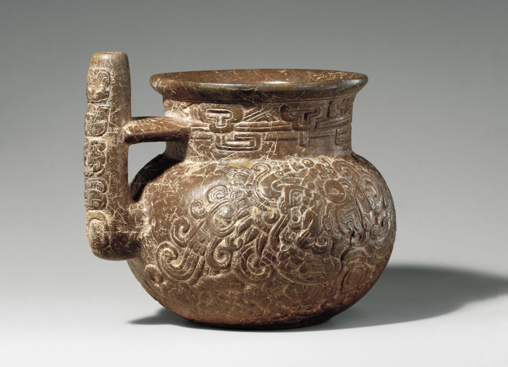 A vessel or mug with an oblong handle is covered in various carvings.