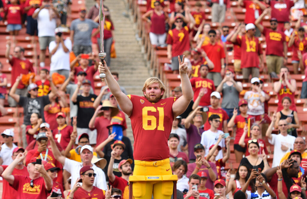 Jake Olson wears his football uniform as he holds up his arms to conduct in front of a crowd of fans, many wearing red USC t shirts.