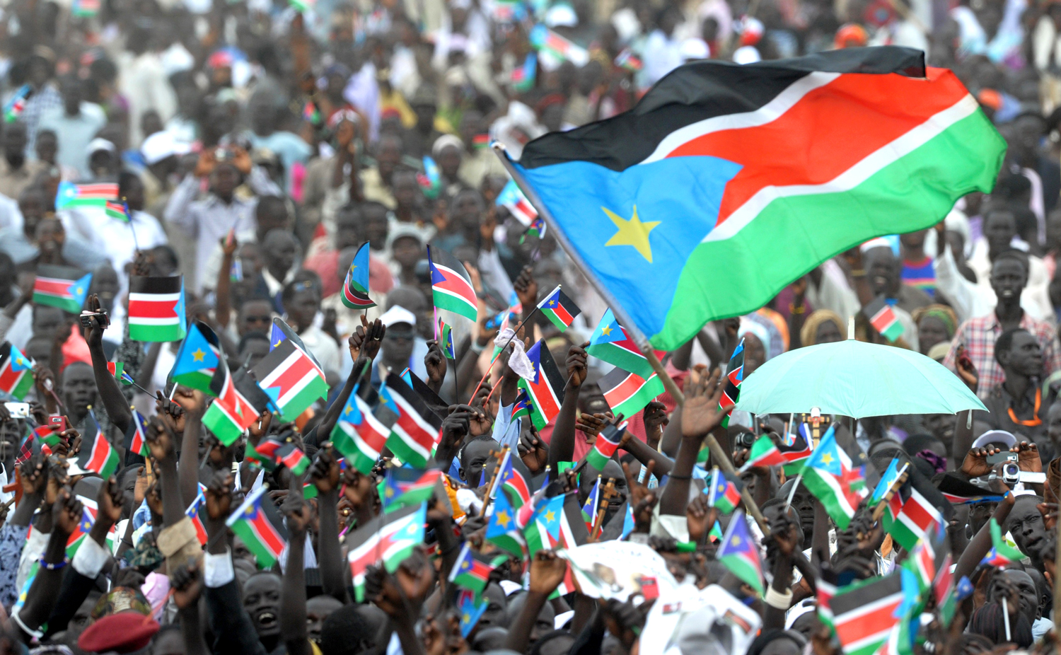 A large crowd of people are gathered and waving South Sudanese flags.