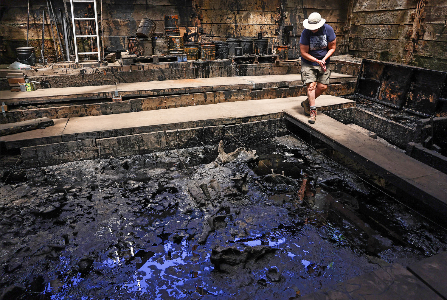 A person wearing a hat walks on the edge of a tar pit that contains visible fossils.