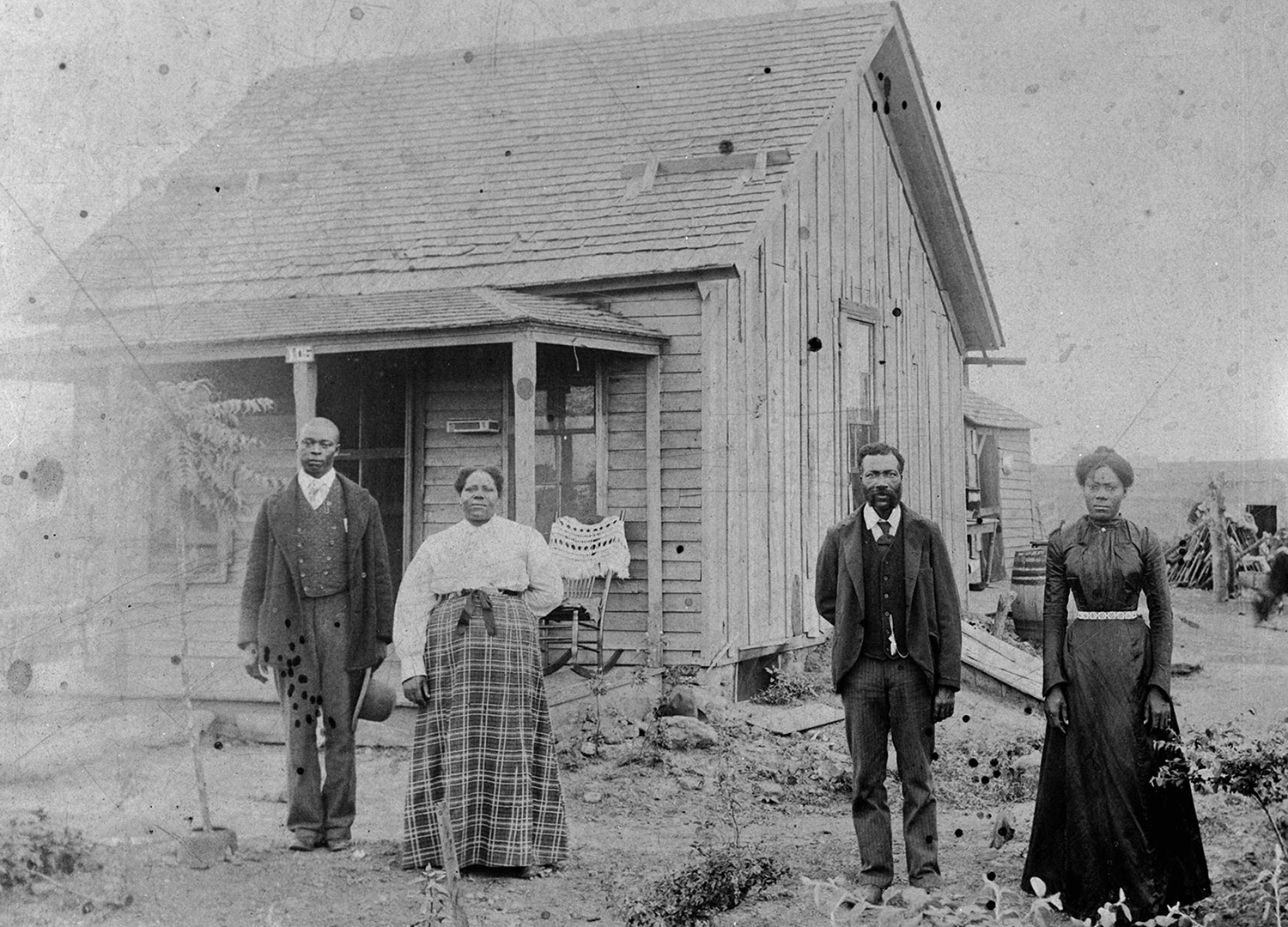 Four Black Americans wearing 19th century clothing stand in front of a small cabin.